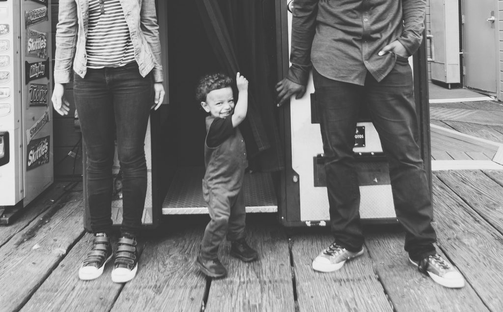 B&W photo of cute little boy playing in a Santa Monica Pier photobooth with his Mom & Dad nearby. Southern California family photographers, Birch Blaze Studios.