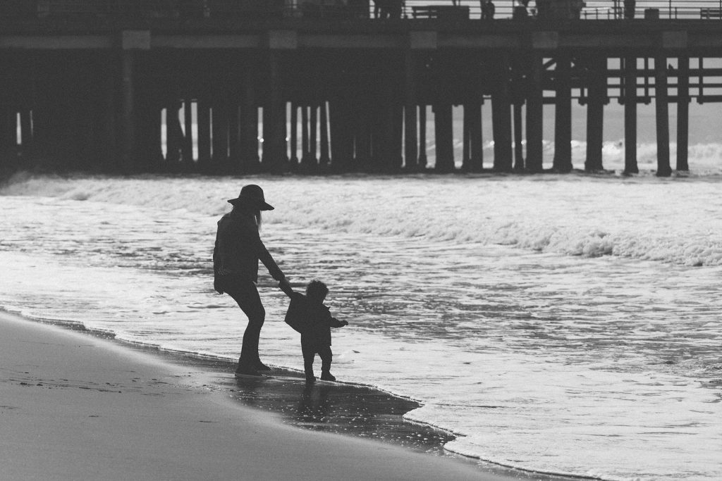 Black & white photo of the silhouettes of a  mother and young son on a southern California beach in front of the Santa Monica Pier. Family photography by Birchblaze.